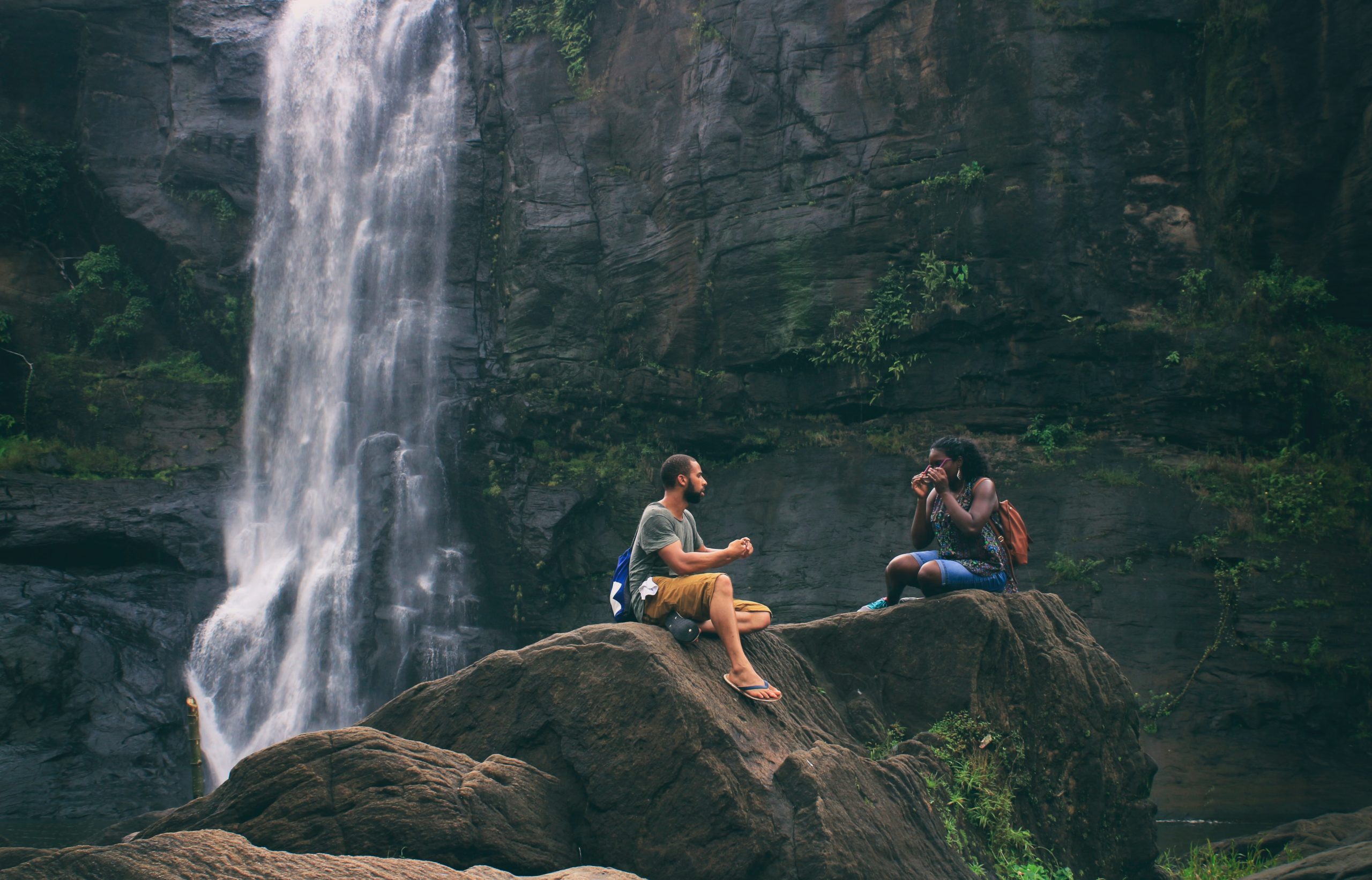 Two people sitting on rocks in front of a waterfall.