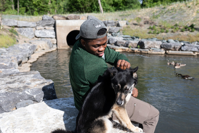 A person wearing a gray baseball hat and green sweater sitting by a river with their dog.